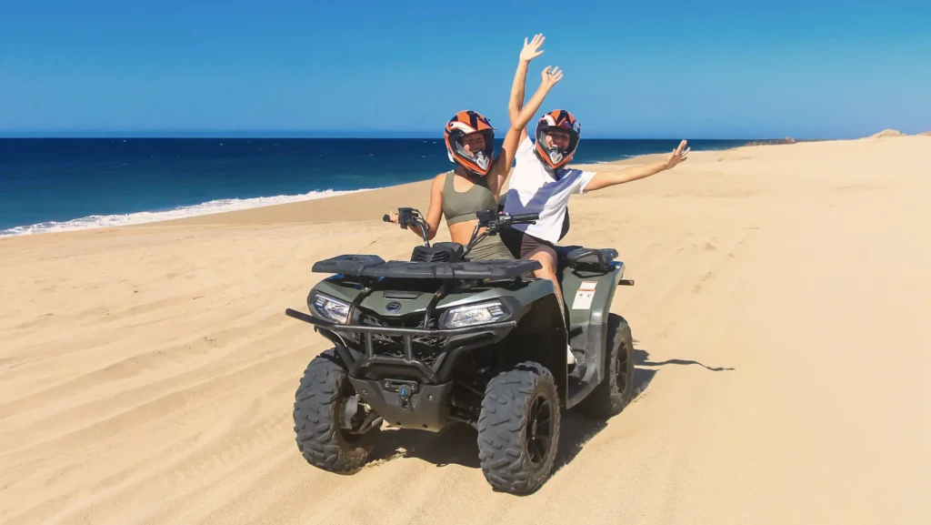 Una pareja usando cascos conduce un vehículo ATV sobre la arena junto al mar, levantando los brazos mientras recorren una playa desértica con el océano al fondo.