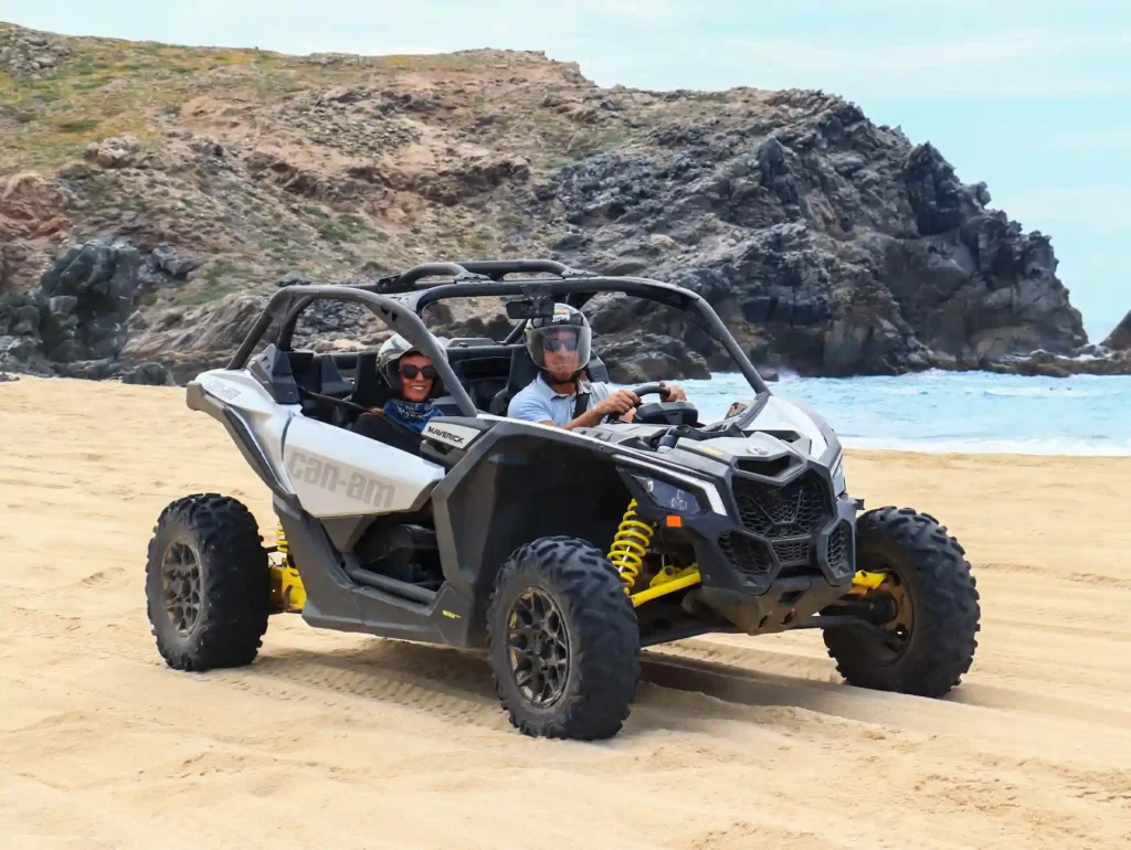 Pareja conduciendo un UTV Maverick X3 sobre la arena junto al mar, con formaciones rocosas al fondo.